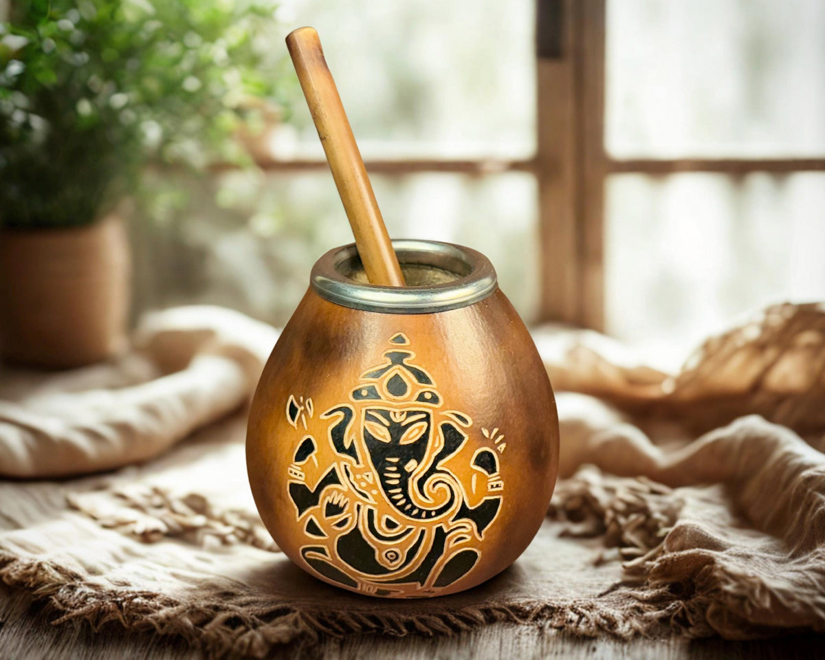 A brass-style mate cup with a wooden straw, decorated with a black Ganesha cutout design, resting on a rustic woven textile with a softly blurred indoor background.
