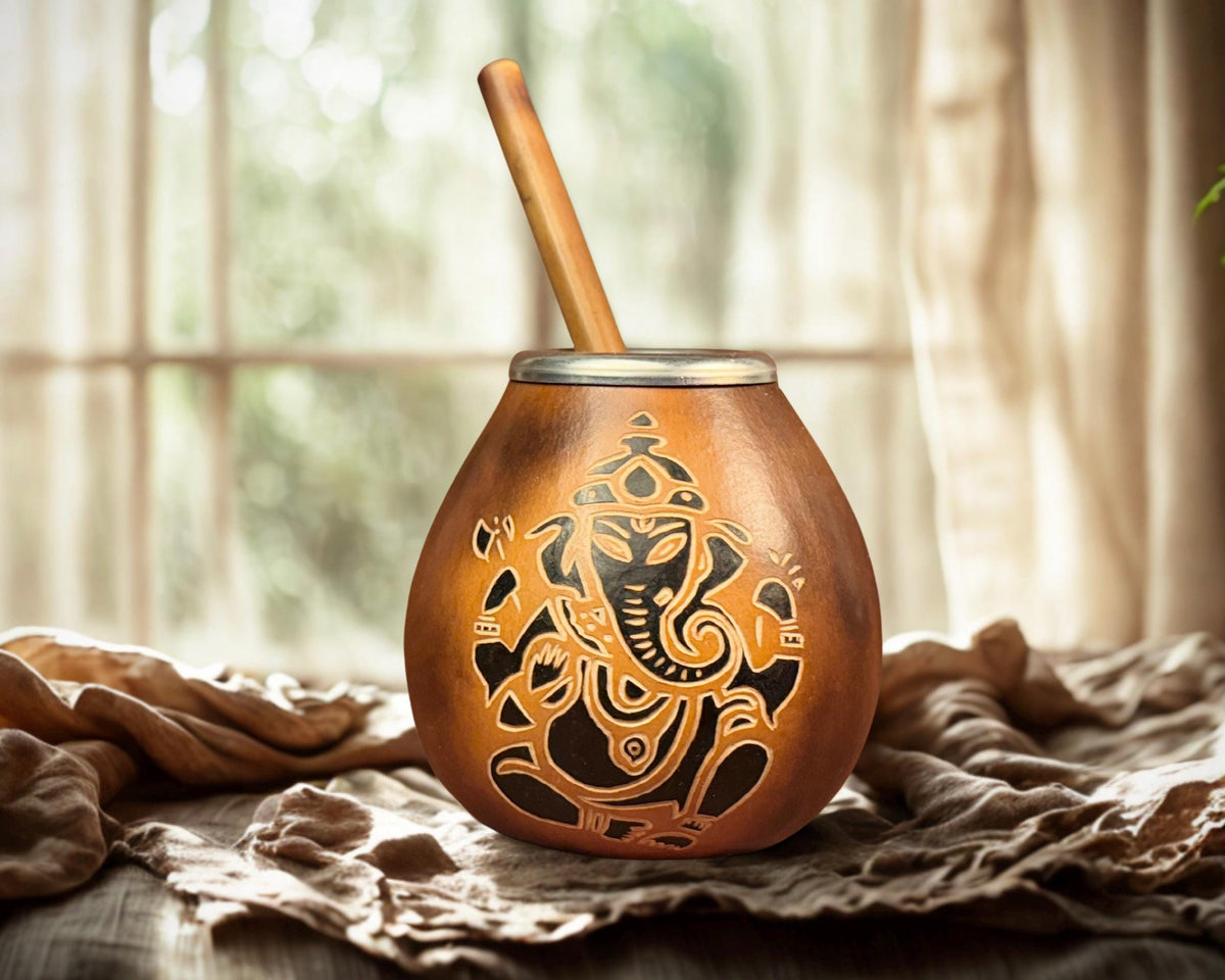 A copper-colored metal mate cup with a Ganesha cutout design and a wooden straw, set on a draped beige cloth in front of a softly blurred window background.