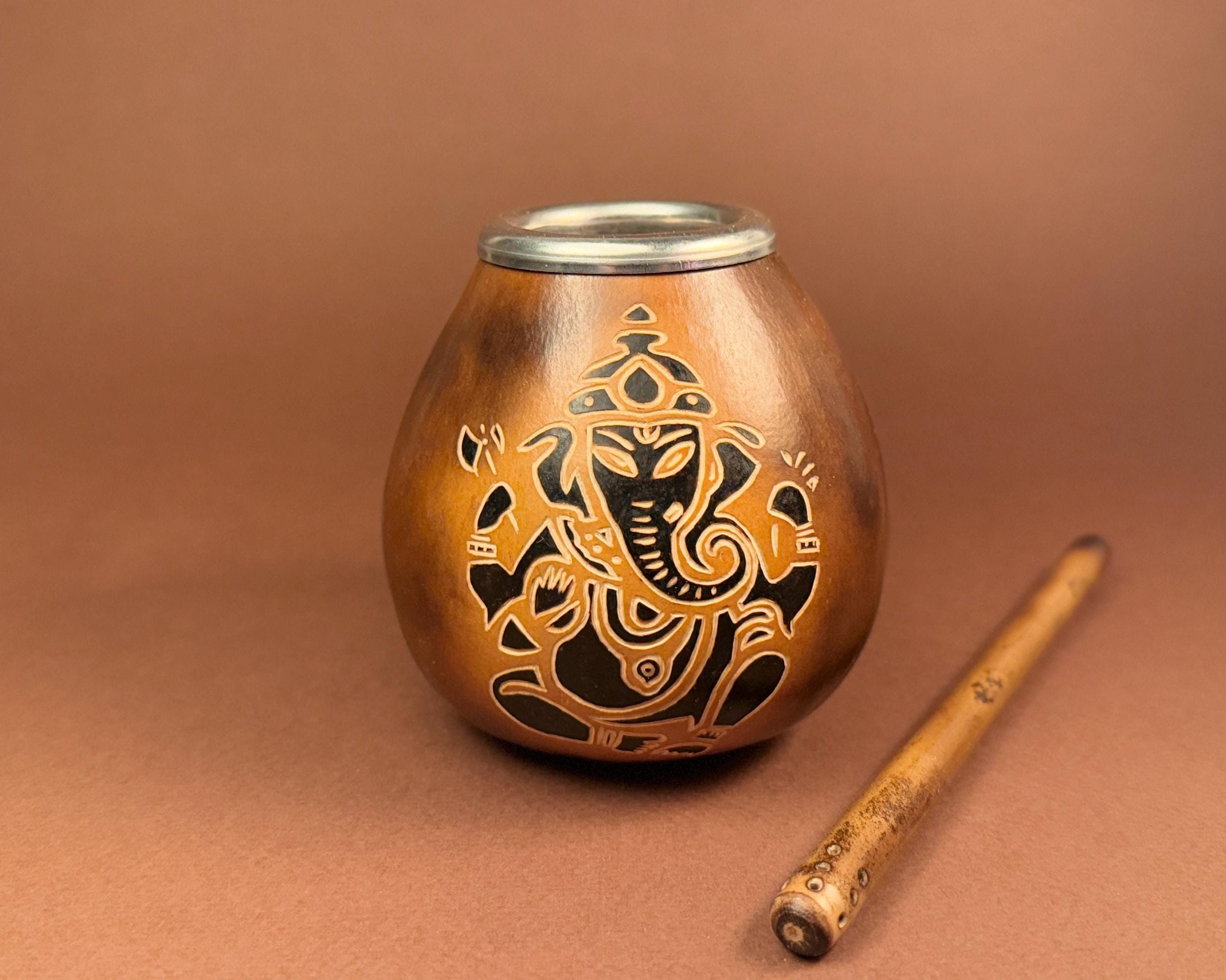 A copper-colored metal shisha container with a Ganesha cutout design and a silver metal rim, shown next to a wooden stick on a brown background