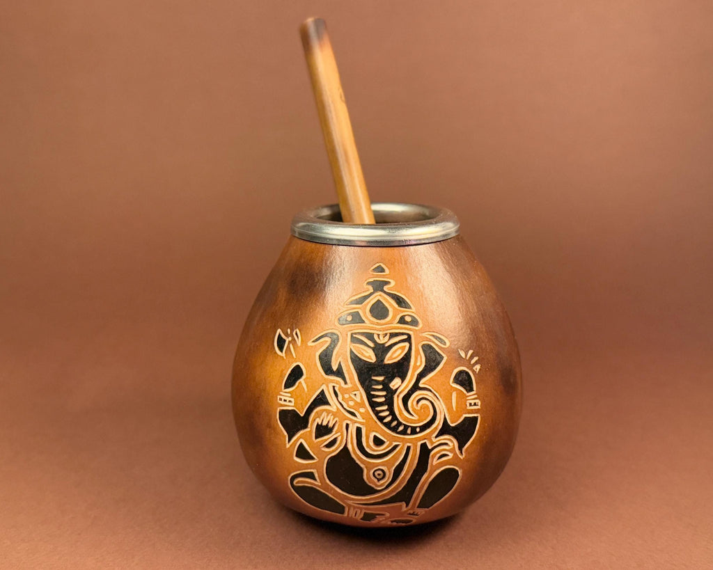 A copper-colored metal mate cup with an engraved Ganesha design and a wooden straw, set against a solid brown background.