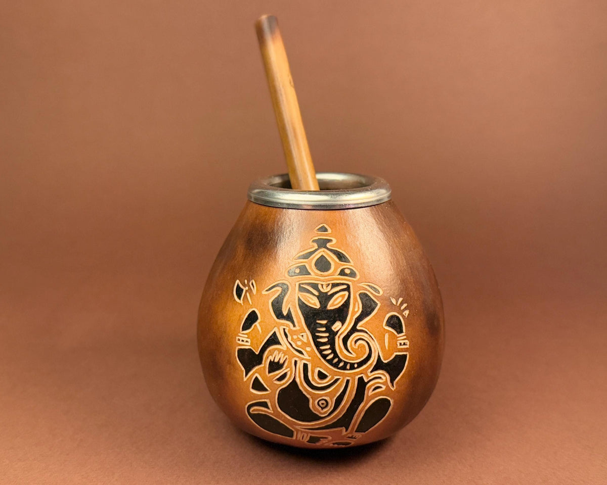 A copper-colored metal mate cup with an engraved Ganesha design and a wooden straw, set against a solid brown background.