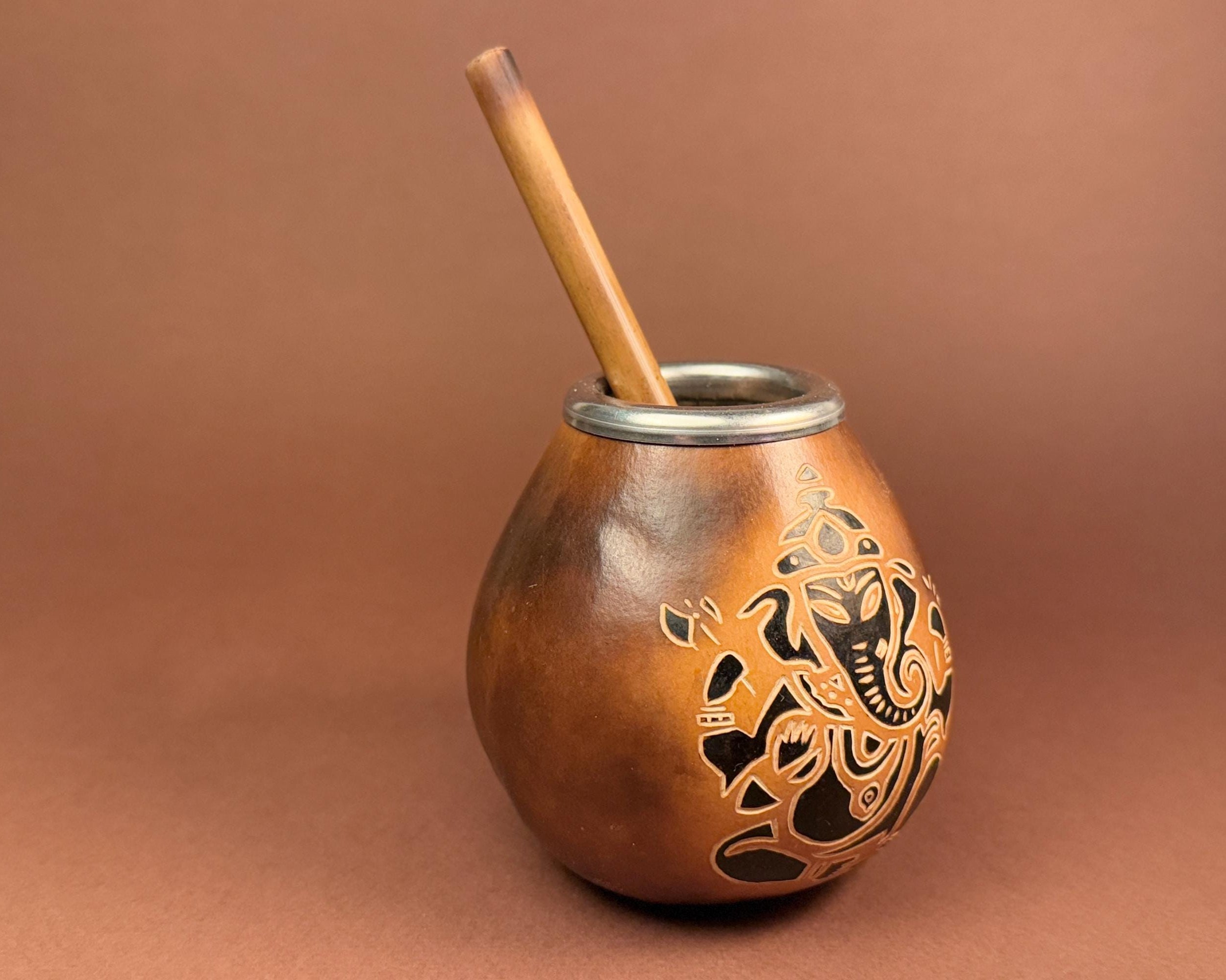 A copper-colored metal mate cup with a wooden straw, featuring a black engraved Guatemalan coat of arms on the front, set against a solid brown background.