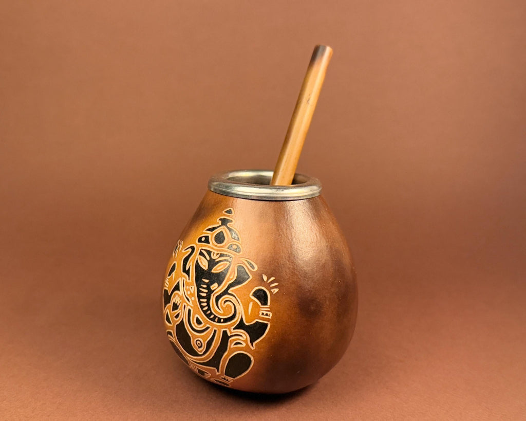 A copper-colored metal mate cup with a silver rim, featuring a black and gold Guatemalan-style tribal graphic on the front, and a wooden straw, set against a solid brown background.