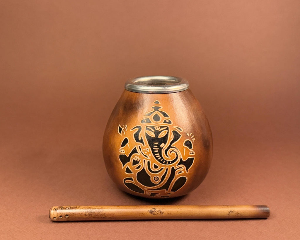 A small metallic copper-toned vessel with a silver rim and an engraved black Ganesha design, shown with a wooden flute on a warm brown background