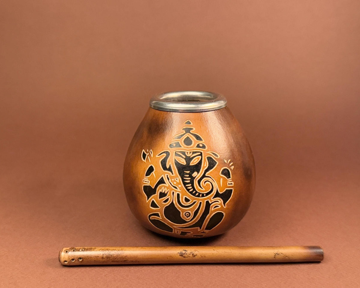 A small metallic copper-toned vessel with a silver rim and an engraved black Ganesha design, shown with a wooden flute on a warm brown background