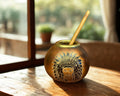 A round brass mate cup with an engraved profile of a figure in a feathered headdress and a light wooden spoon inside, set on a rustic wooden table in warm natural light.