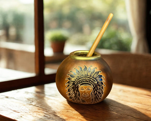 A round brass mate cup with an engraved profile of a figure in a feathered headdress and a light wooden spoon inside, set on a rustic wooden table in warm natural light.