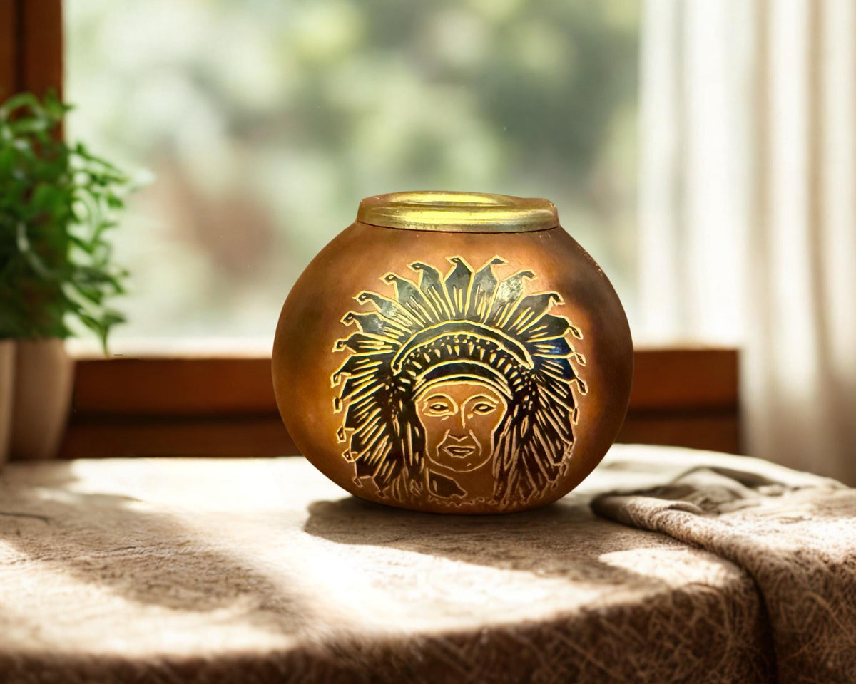 A round metallic bronze decorative urn with an engraved profile of a face wearing a feathered headdress, displayed on a draped tablecloth in a sunlit room near a window.