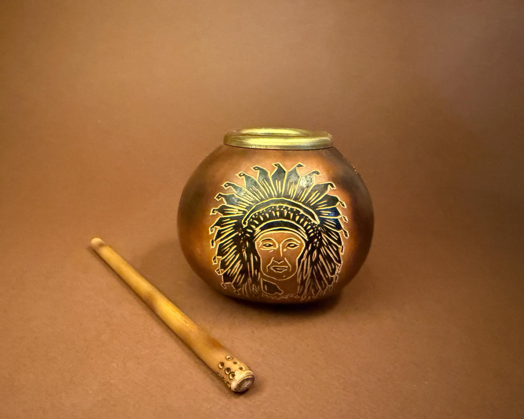 A small round metallic tin or candle holder with a lid, featuring an engraved Native American headdress design, shown on a brown background next to a wooden stick.