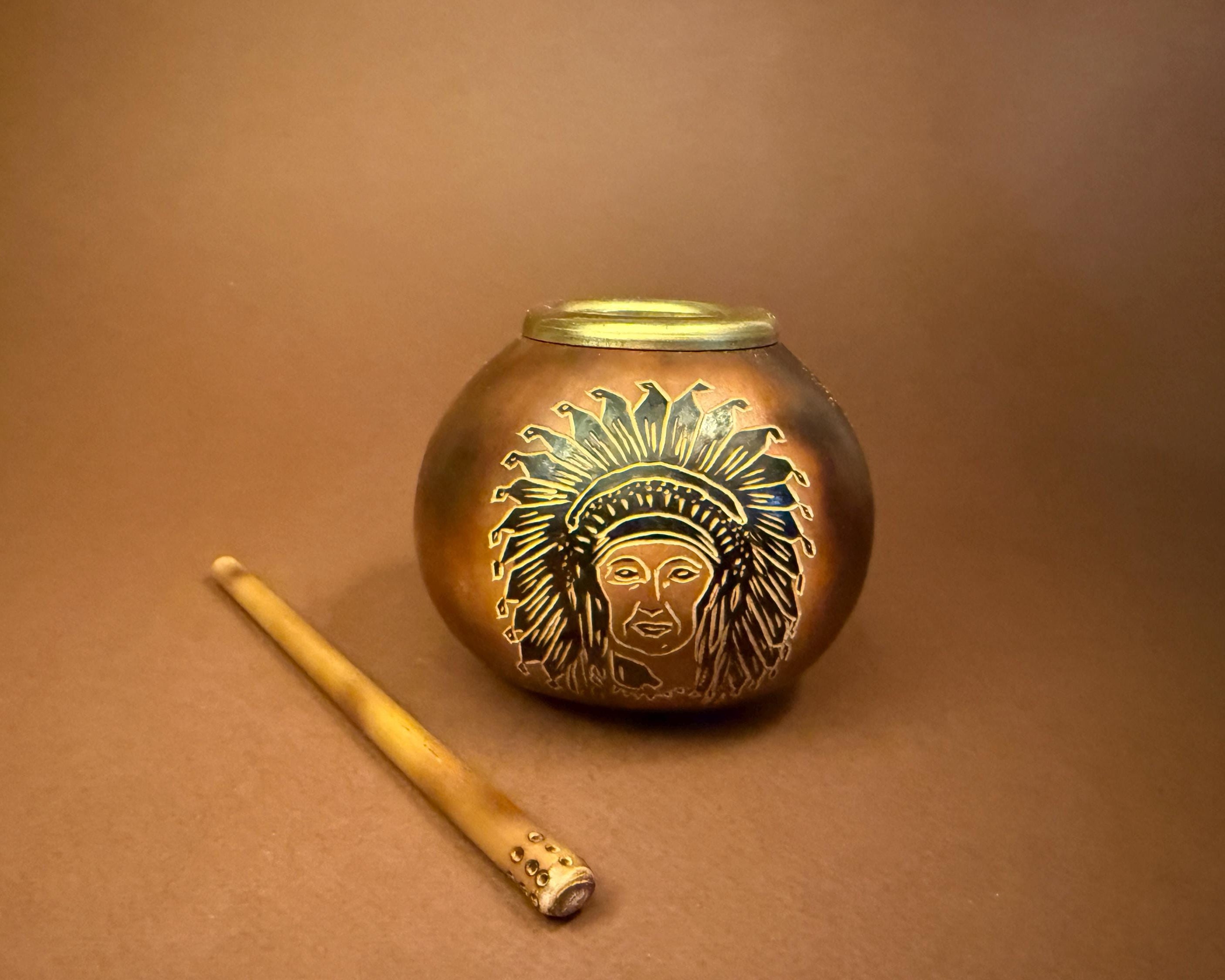 A small round metallic tin or candle holder with a lid, featuring an engraved Native American headdress design, shown on a brown background next to a wooden stick.