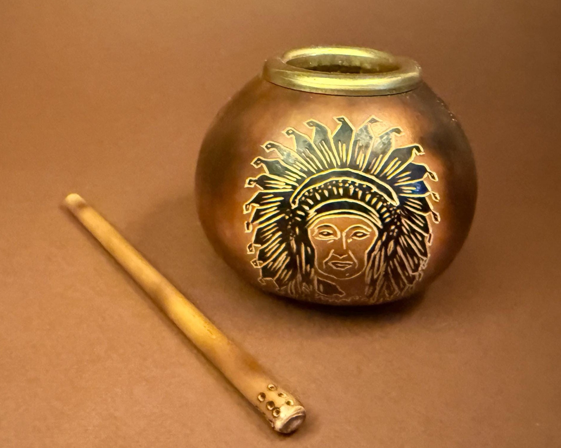 A small round metallic pot with a gold-toned rim and an engraved face wearing a feathered headdress, shown on a brown background next to a thin wooden stick.