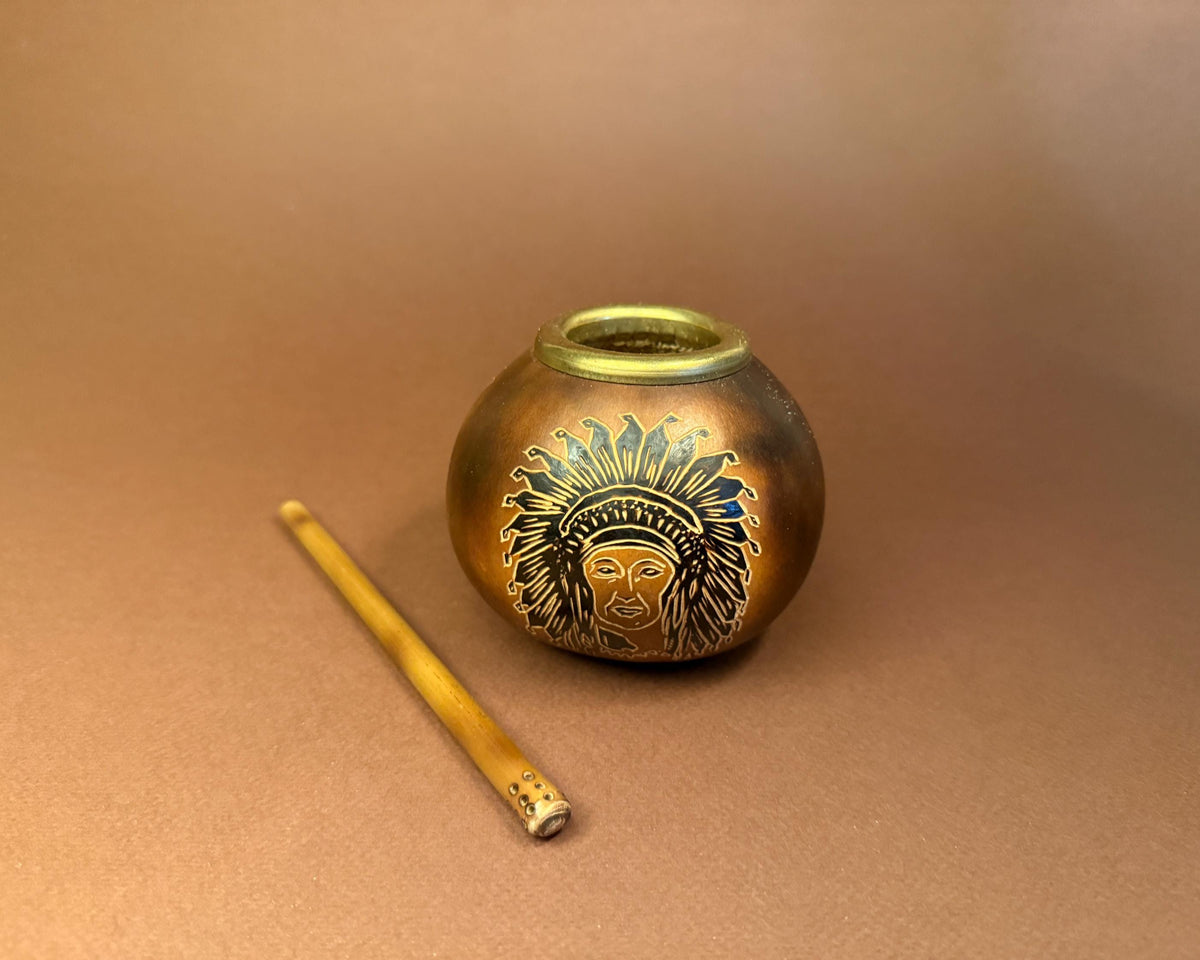 A small round metallic vessel with an engraved face wearing a feathered headdress, shown on a warm brown background next to a thin yellow rod.