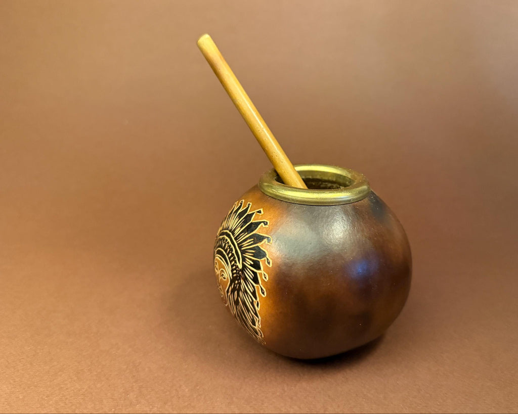 A small handcrafted ceramic mate pot with a wooden straw, featuring a metallic bronze and gold finish with a black and gold sun design on the side, set against a solid brown background.