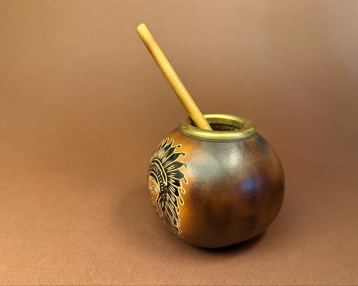 A small handcrafted ceramic mate pot with a wooden straw, featuring a metallic bronze and gold finish with a black and gold sun design on the side, set against a solid brown background.