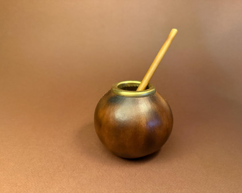 A small round brown metal mate pot with a brass rim, containing a light tan wooden straw, set against a solid brown background.