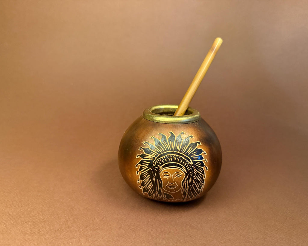 A small round metallic cup with an engraved face wearing a feathered headdress, shown on a warm brown background with a light wooden straw inserted.