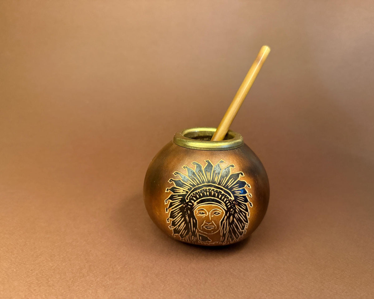 A small round metallic cup with an engraved face wearing a feathered headdress, shown on a warm brown background with a light wooden straw inserted.