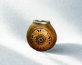A small handcrafted clay pot with a metallic rim and intricate black geometric patterns on the front, set against a textured white background with soft shadows.
