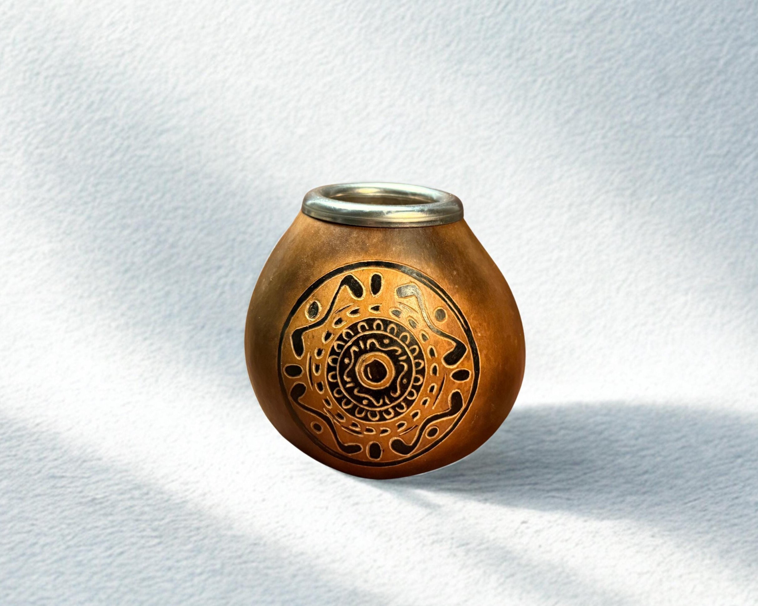 A small handcrafted clay pot with a metallic rim and intricate black geometric patterns on the front, set against a textured white background with soft shadows.