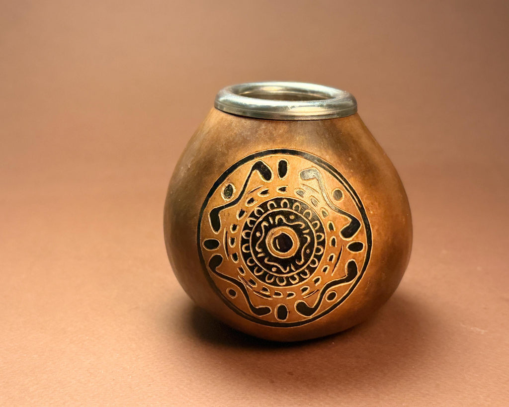 A small copper gourd or mate pot with a silver metal rim, featuring a black hand-painted circular geometric pattern on the front, set against a solid brown background.