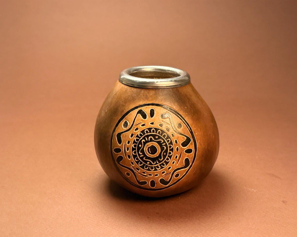 A small handcrafted wooden goblet or cup with a polished copper-brown finish, featuring an intricate circular cutout pattern on the front and a silver metal rim, set against a solid warm brown background