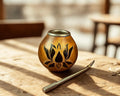 cup with a black lotus floral design, placed on a rustic wooden table next to a metal straw in a warm, sunlit setting