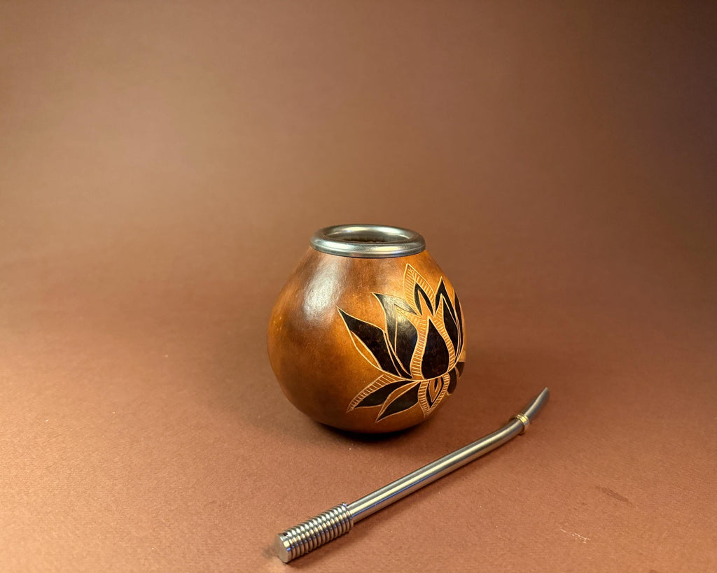 A small handcrafted wooden gourd pipe with a silver metal rim and an engraved lotus flower design, shown next to a silver metal bombilla on a brown background