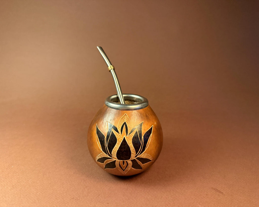 A small copper-colored metal mate cup with a black lotus flower inlay on the front and a silver metal straw, photographed against a smooth warm brown gradient background