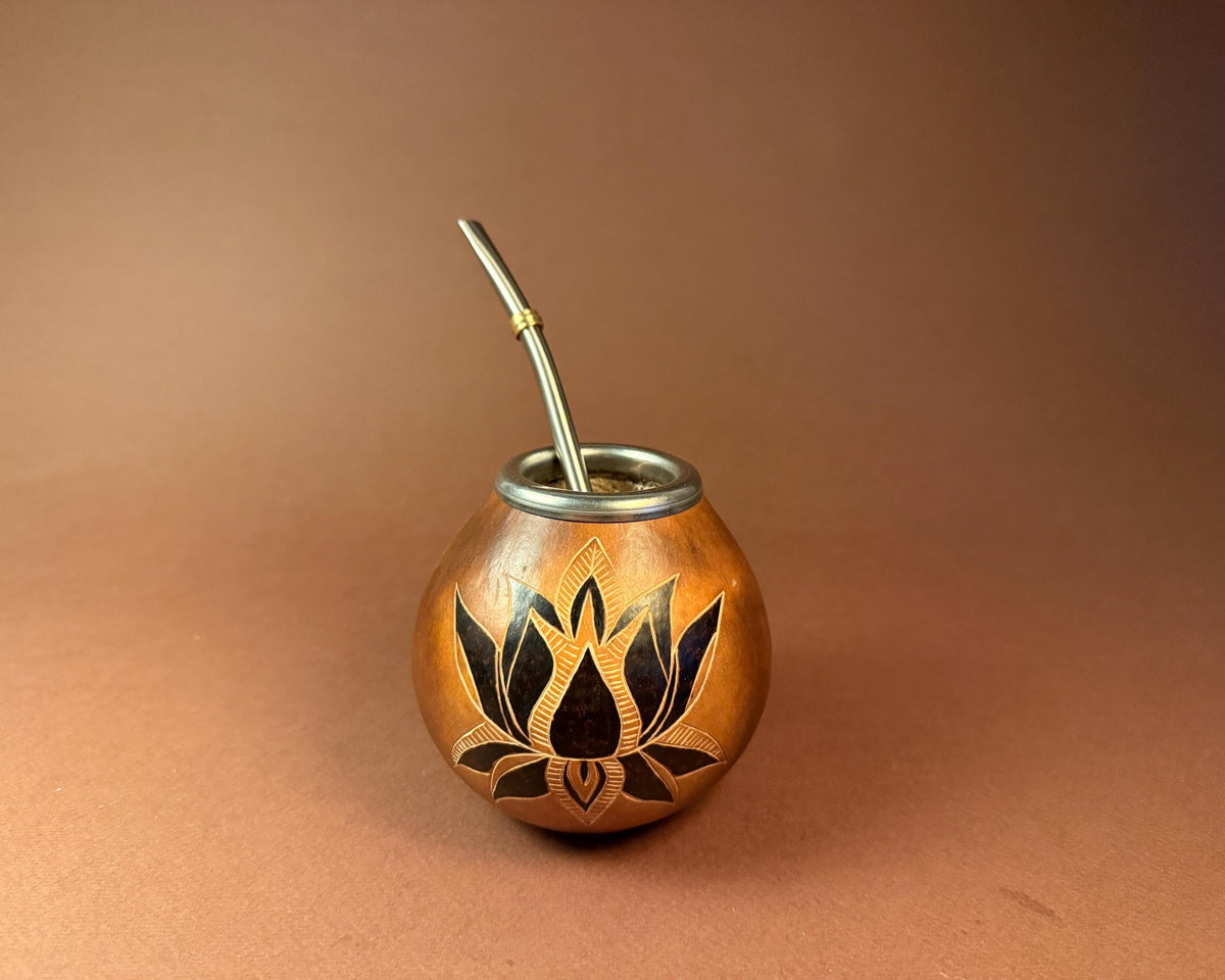 A small copper-colored metal mate cup with a black lotus flower inlay on the front and a silver metal straw, photographed against a smooth warm brown gradient background