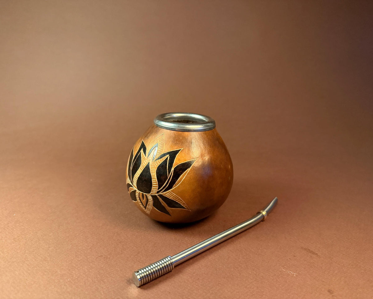 A small copper-toned gourd with black leaf inlay and a silver metal rim, shown on a warm brown studio background with a silver metal bombilla resting in front.