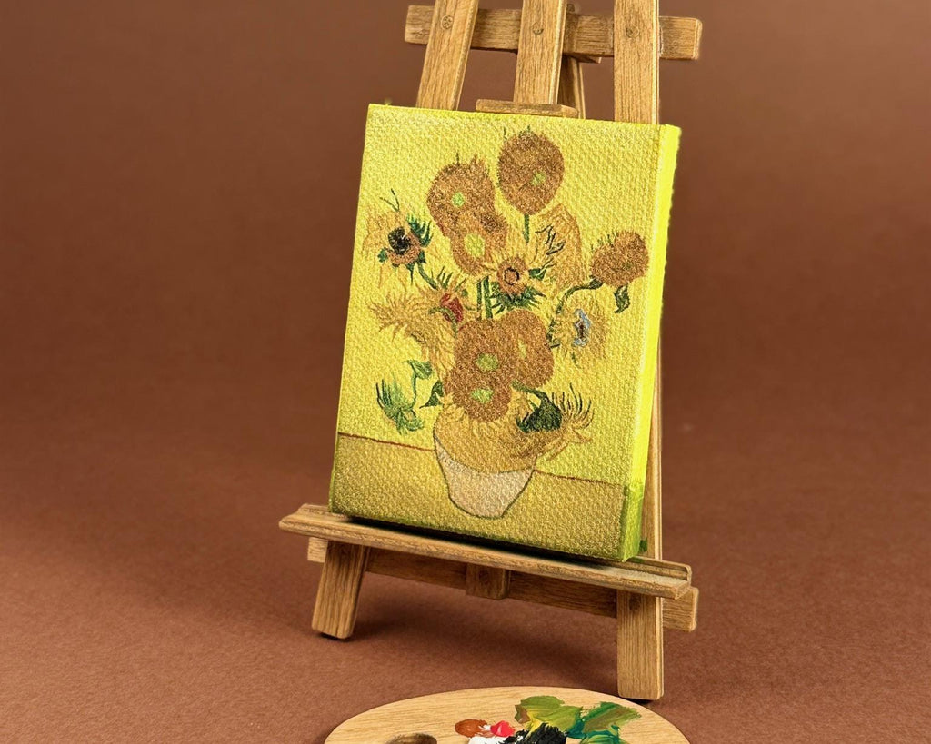 A miniature canvas easel holding a small canvas print of Van Gogh's sunflowers, with a matching miniature wooden paint palette in front, set against a solid brown background