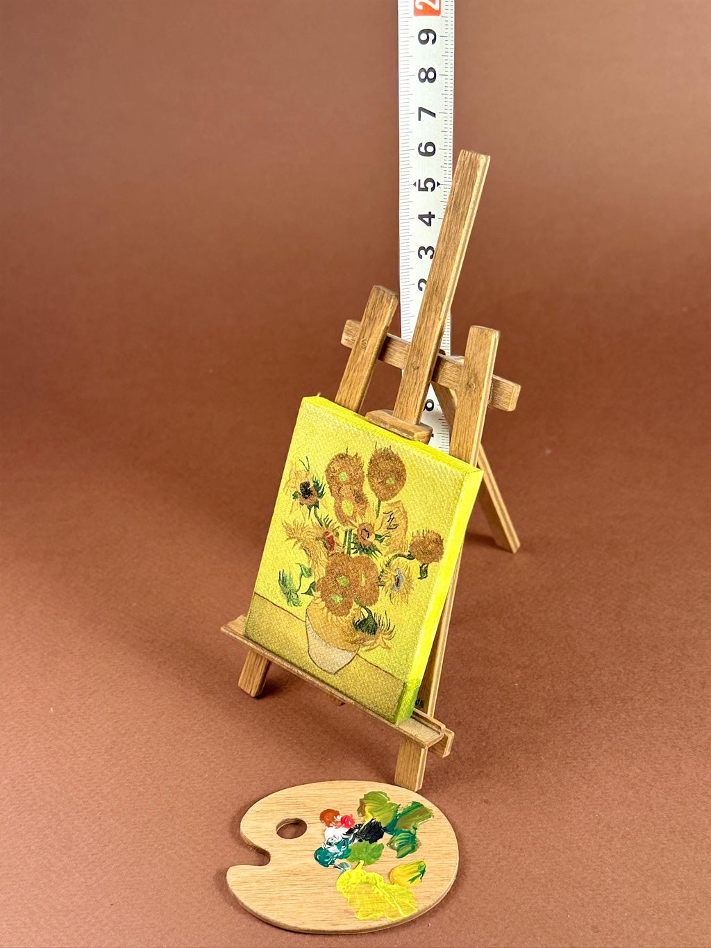 A miniature wooden artist easel holding a small canvas painting of sunflowers, with a small wooden paint palette in front and a measuring tape standing behind it on a brown studio background.