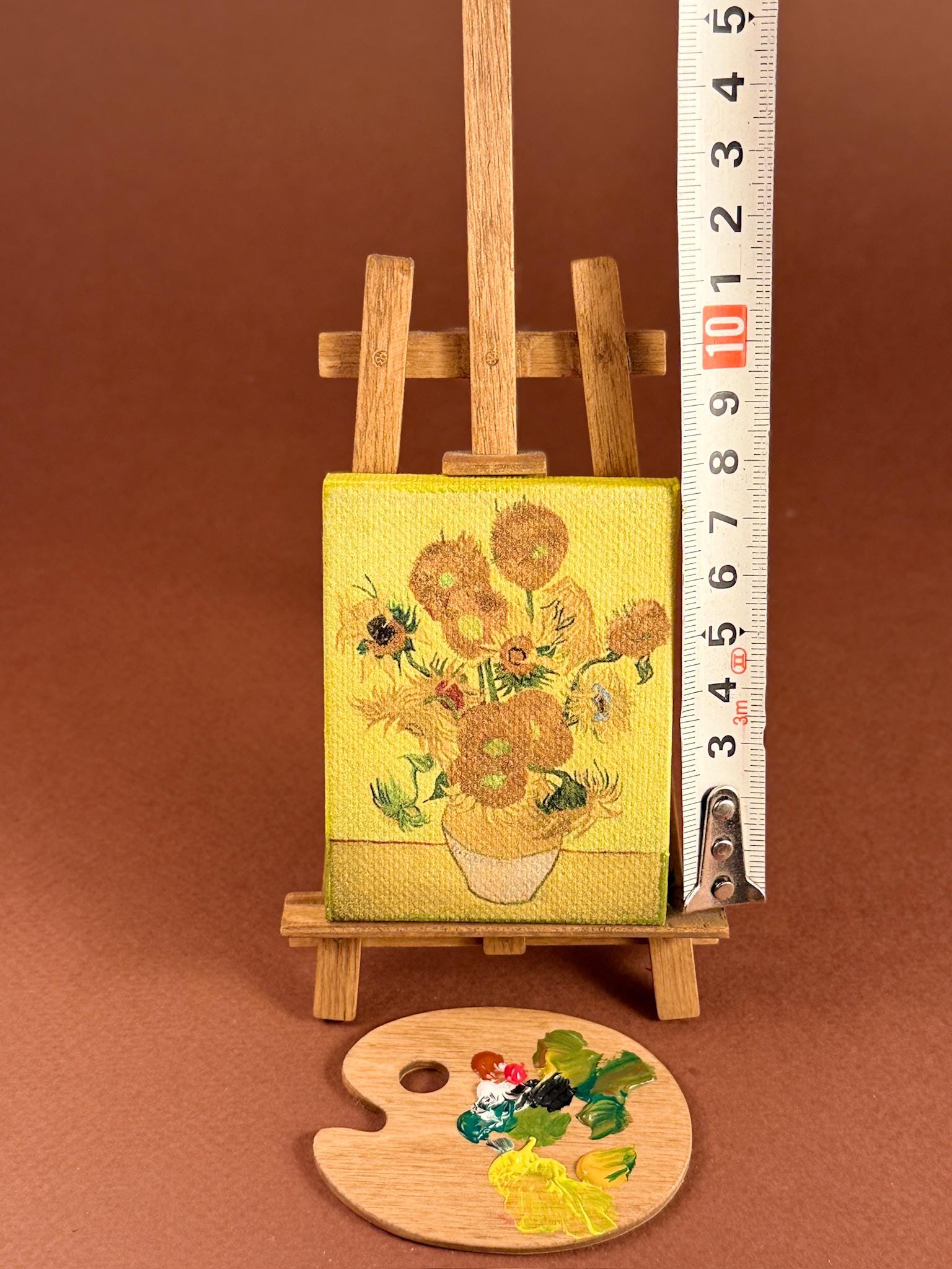 A miniature canvas easel holding a small painted bouquet of yellow flowers on yellow fabric, shown next to a measuring tape and a wooden paint palette on a brown background.