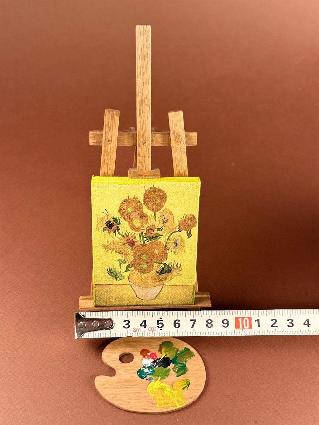A miniature wooden easel holding a small yellow canvas painting of sunflowers, shown with a metal ruler and a small wooden paint palette on a brown background.