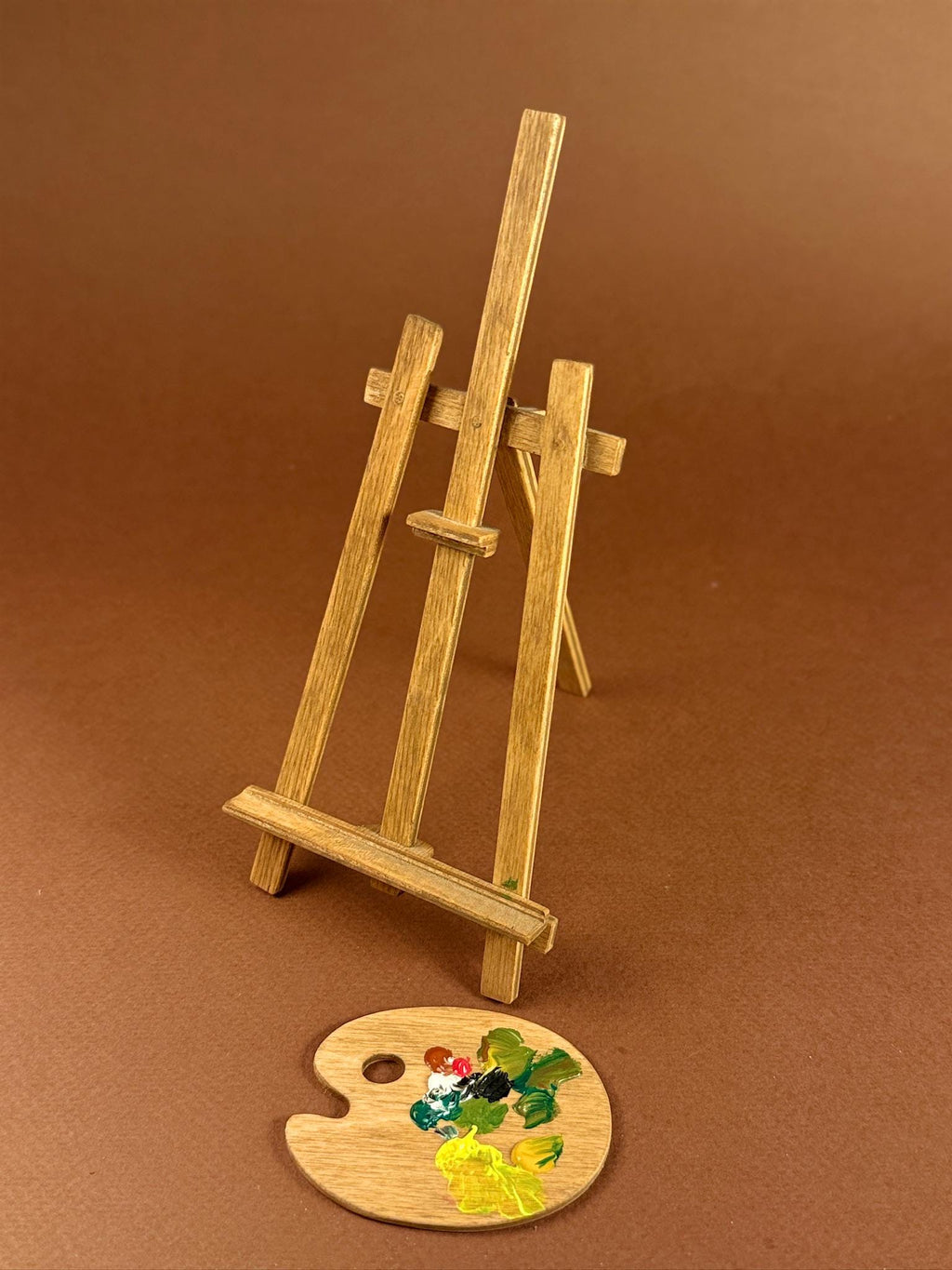 A miniature wooden artist easel stand with a small round wooden paint palette resting in front, set against a solid brown background.