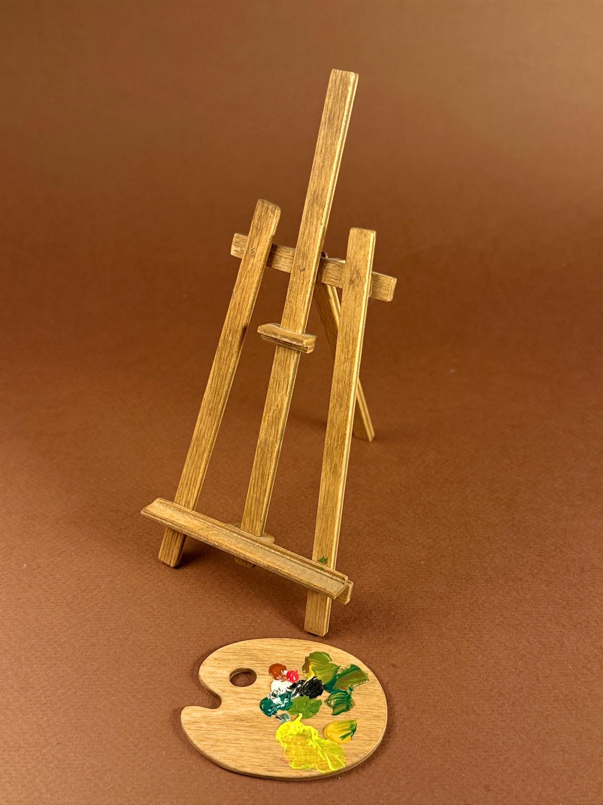 A miniature wooden artist easel stand with a small round wooden paint palette resting in front, set against a solid brown background.
