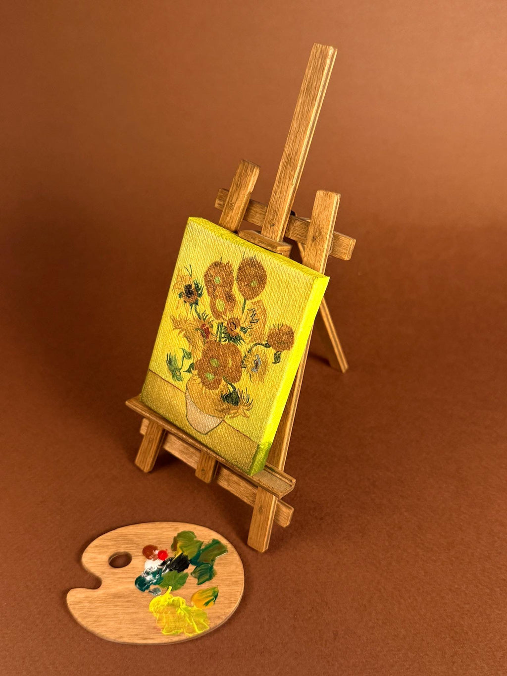 A miniature wooden easel holding a small canvas print of a yellow floral painting, with a matching miniature paint palette on a brown studio background.