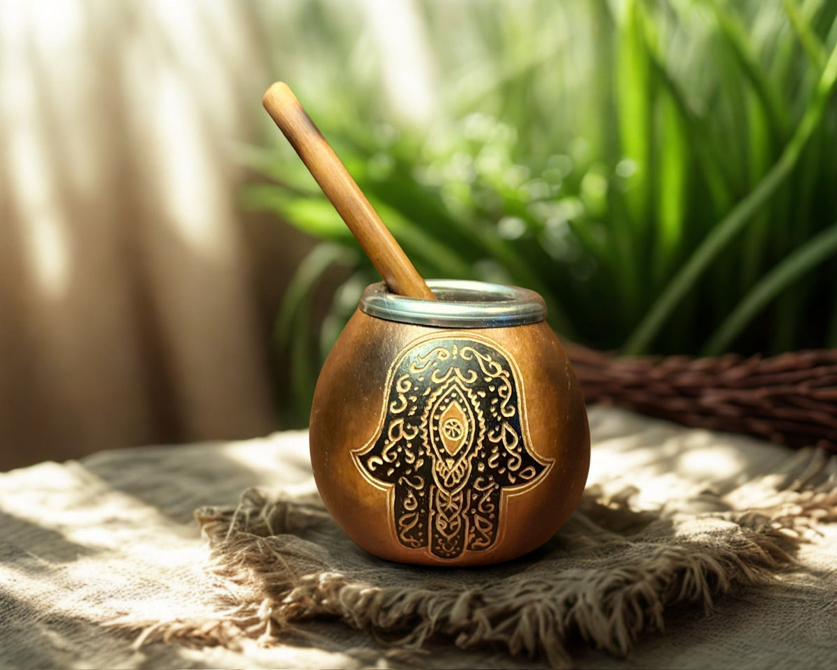 A small round copper or brass mug with a wooden spoon, decorated with a black Hamsa hand motif, resting on a frayed beige cloth with a blurred background of green plants and warm light.
