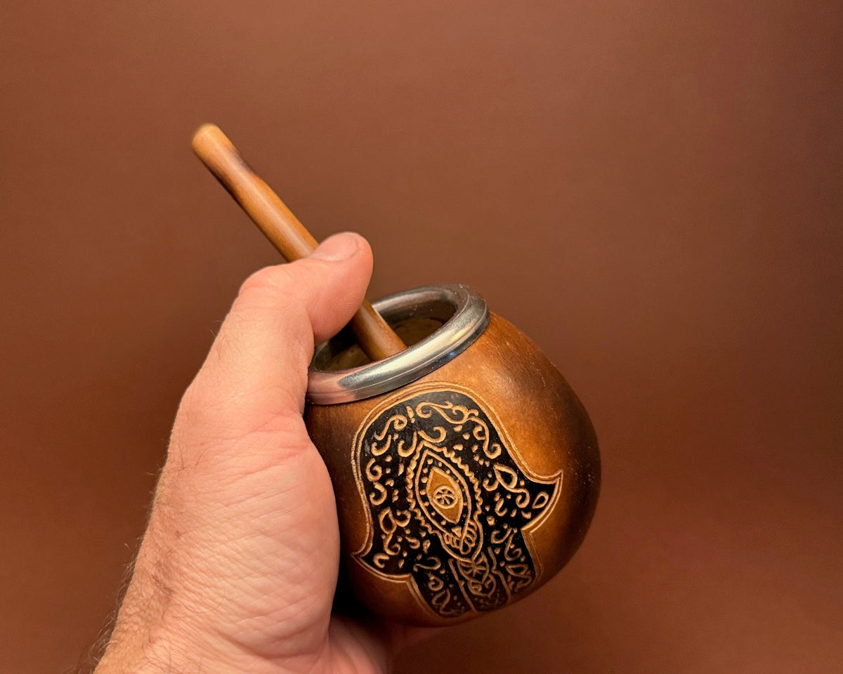 A hand holding a small brown wooden or metal mate cup with a wooden spoon, decorated with a black hamsa hand design, against a solid brown background