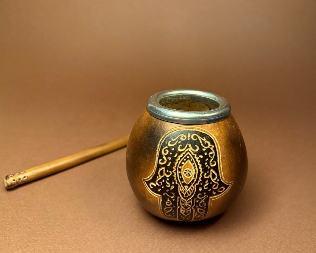 A small round metal container with a decorative black and gold hamsa hand design on the front, shown on a brown background next to a wooden stick