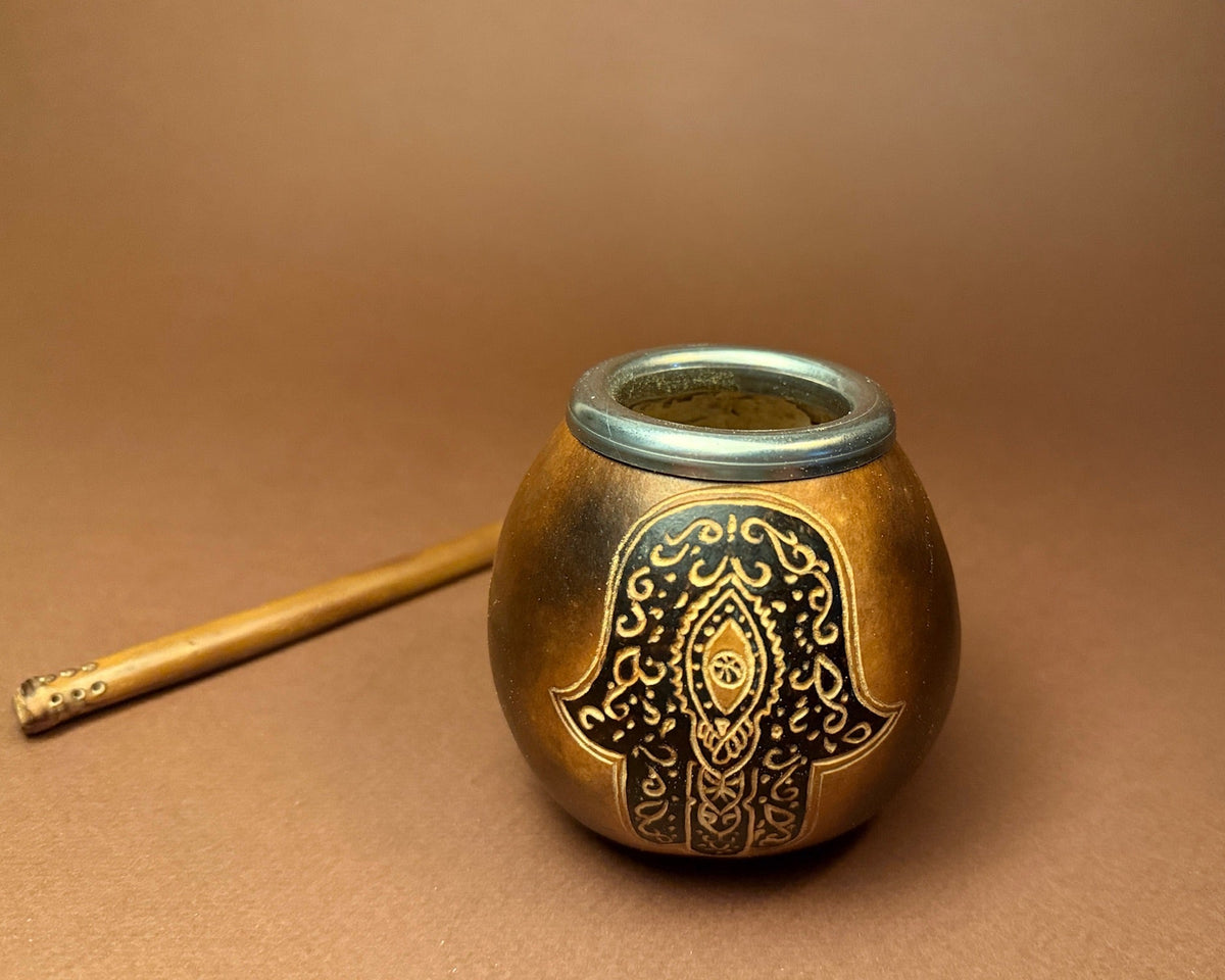 A small round metal container with a decorative black and gold hamsa hand design on the front, shown on a brown background next to a wooden stick