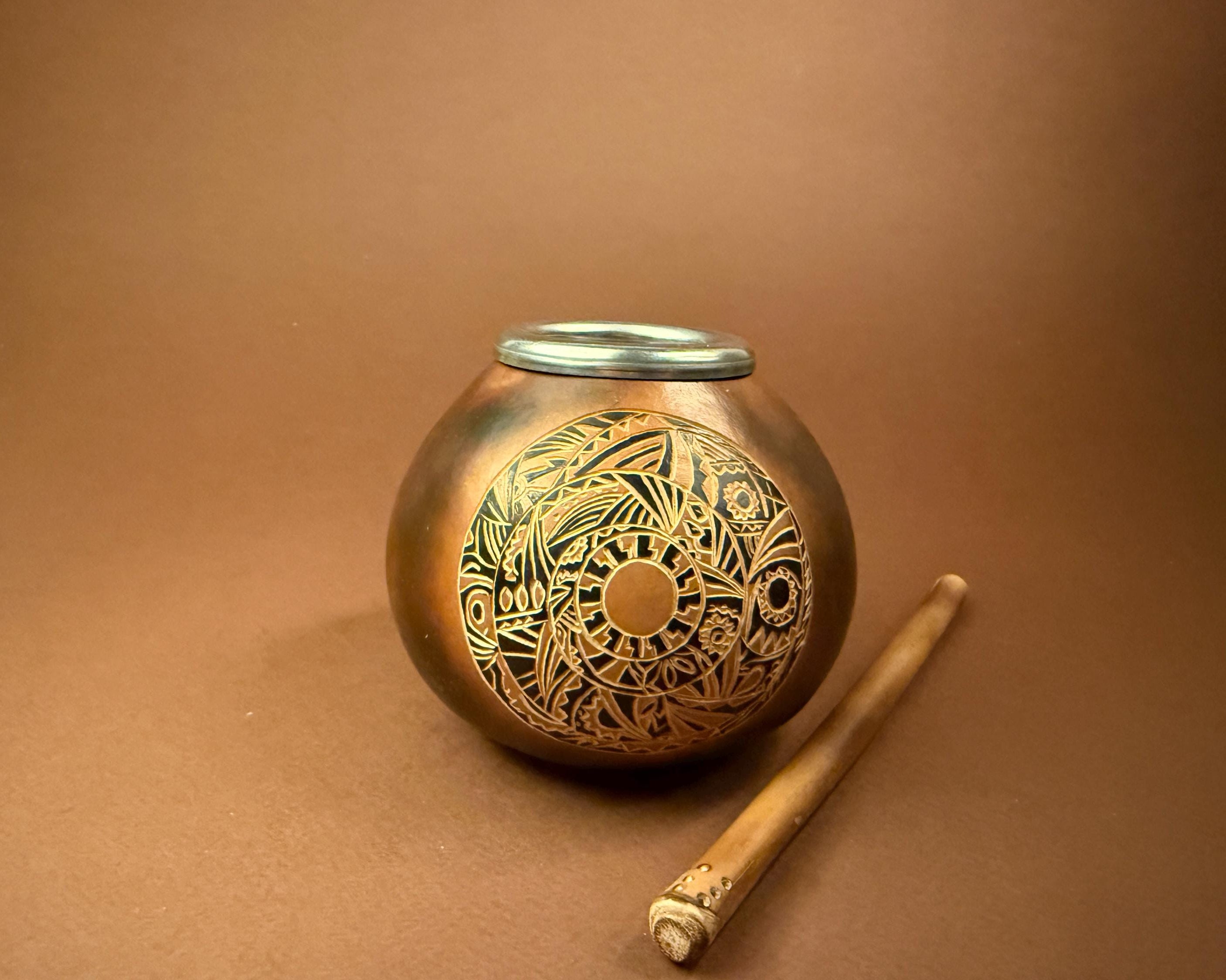 A round bronze-colored metal tin with a lid, decorated with an intricate circular cutout pattern on the front, shown next to a small wooden stick on a brown background.