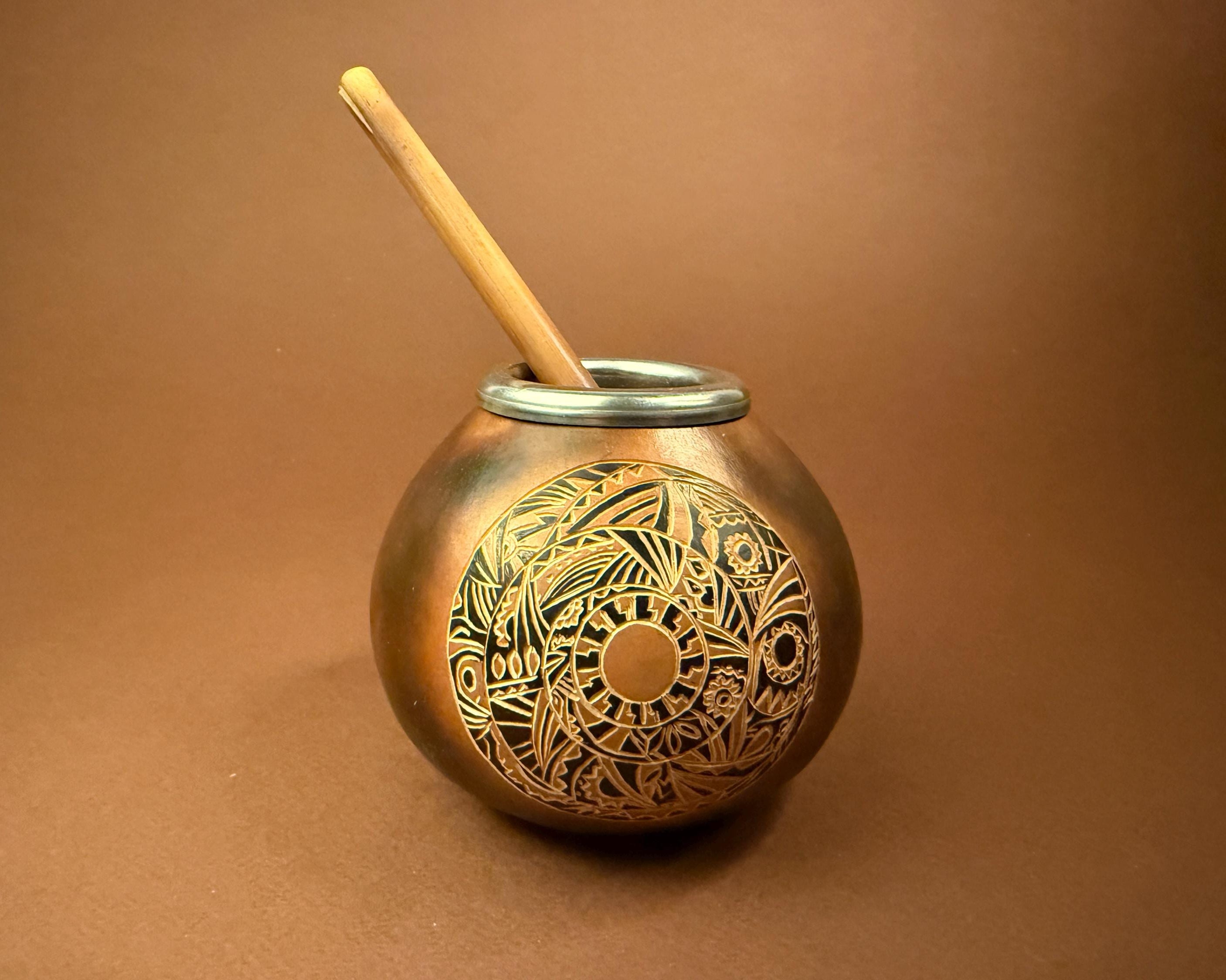 A round copper-colored metal mate pot with an intricate engraved circular pattern on the front, featuring a silver metal rim and a light wood spoon resting inside, set against a solid brown background