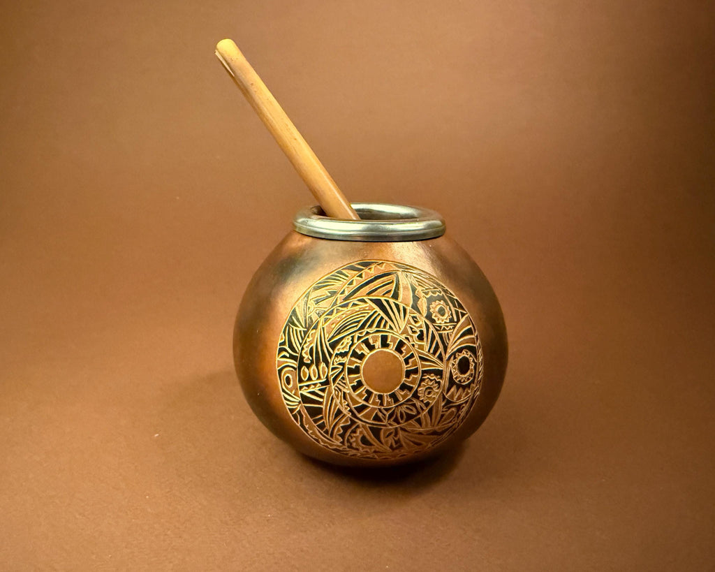 A round copper-colored metal mate cup with an intricate engraved circular pattern on the front, featuring a light wooden straw resting inside, set against a solid brown background.