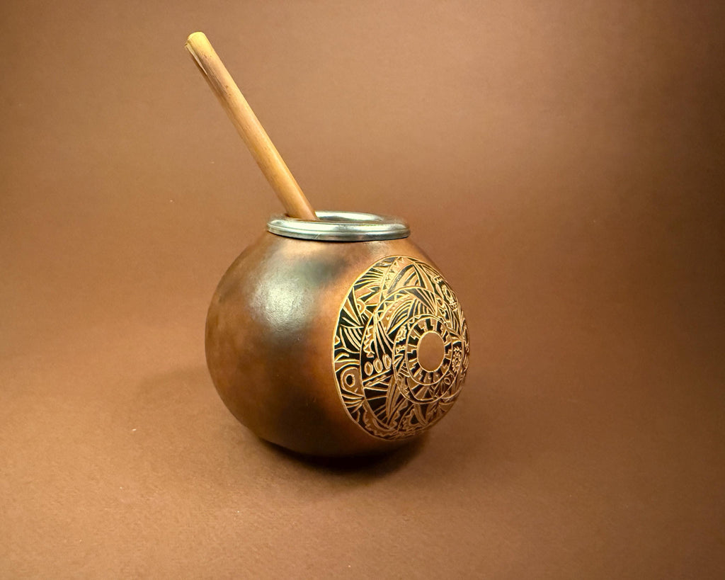 A handmade copper mate pot with an engraved circular design on the side, featuring a silver metal rim and a wooden straw, set against a solid brown background