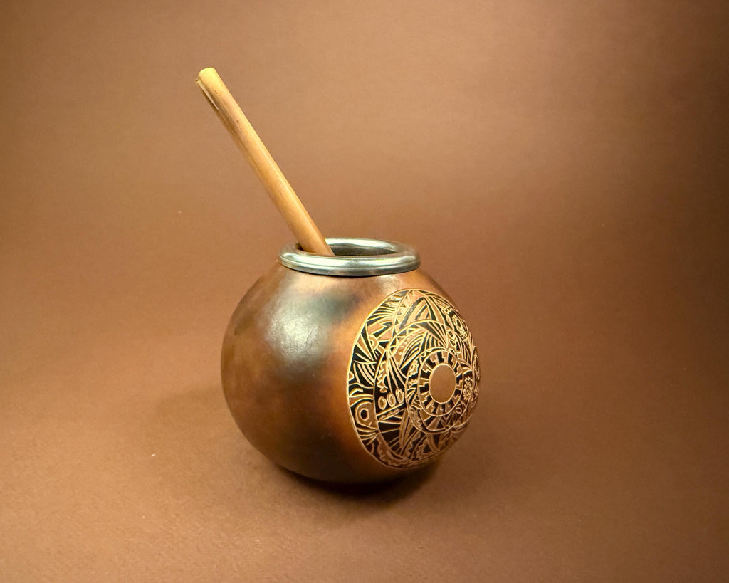 A traditional copper mate pot with an engraved circular pattern on the front, featuring a silver metal rim and a light wood yerba mate straw, set against a solid brown background