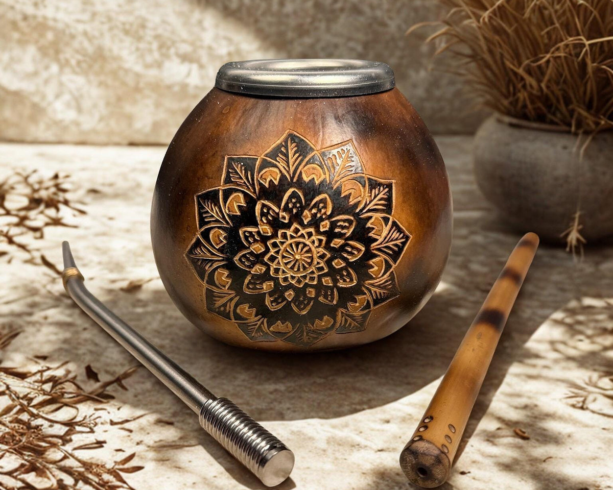 A brown wooden mate pot with a black mandala design, shown with a metal bombilla and a bamboo stem on a stone surface with dried botanicals.