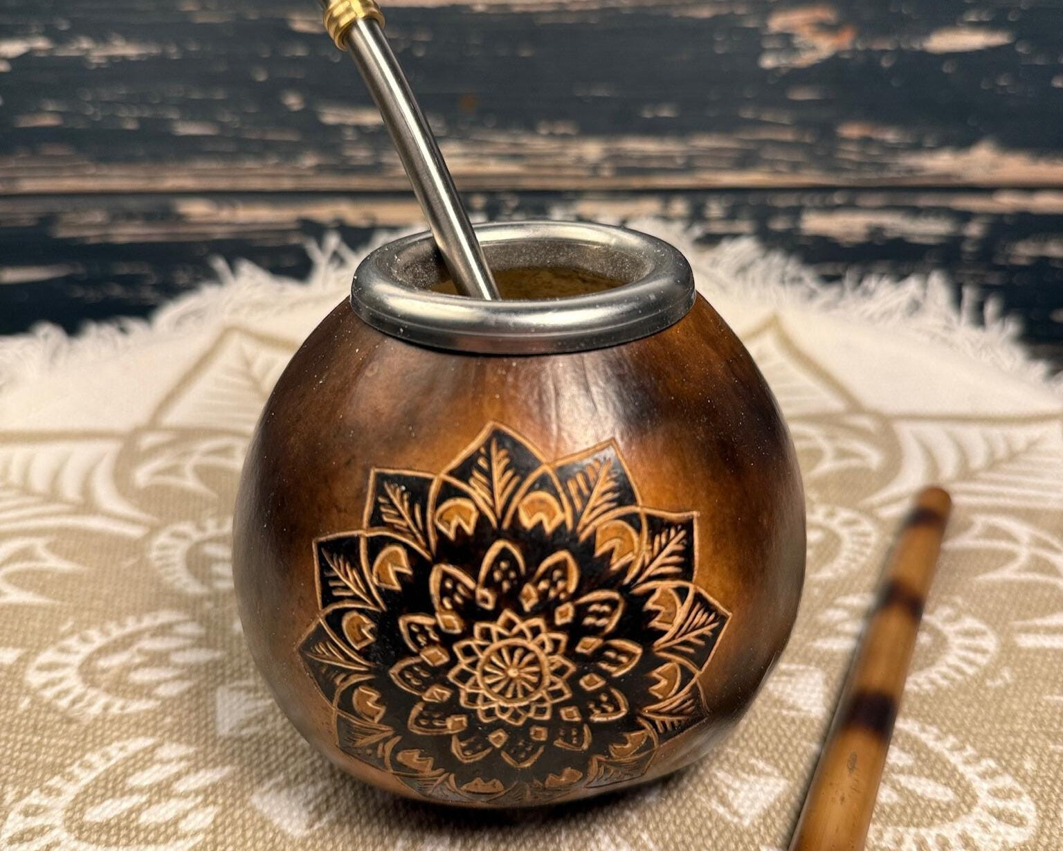 A brown wooden mate pot with an engraved black and gold floral mandala design, featuring a silver metal bombilla straw, set on a beige patterned textile against a dark rustic wooden background.