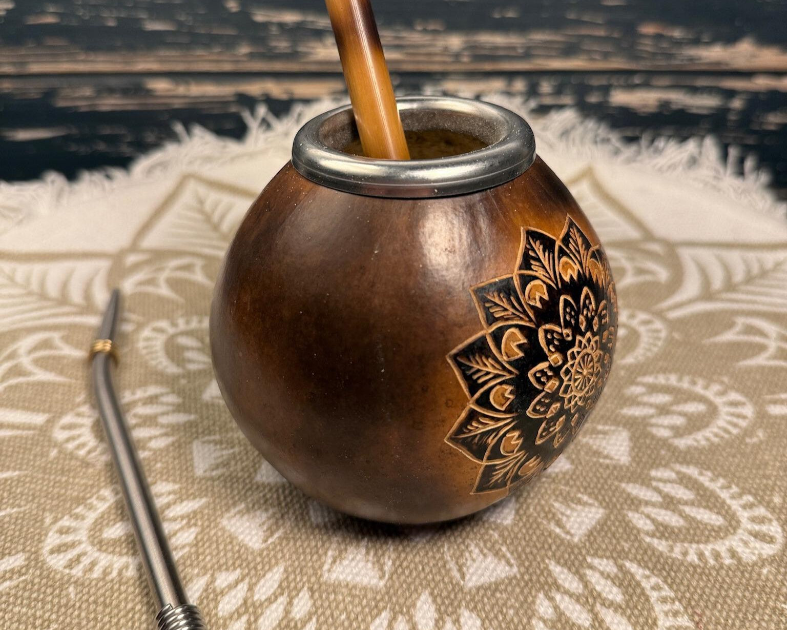 A brown wooden mate bomb with a black floral mandala design on the side, containing a wooden mate straw, shown with a metal bombilla and tenpillo on a patterned beige textile.