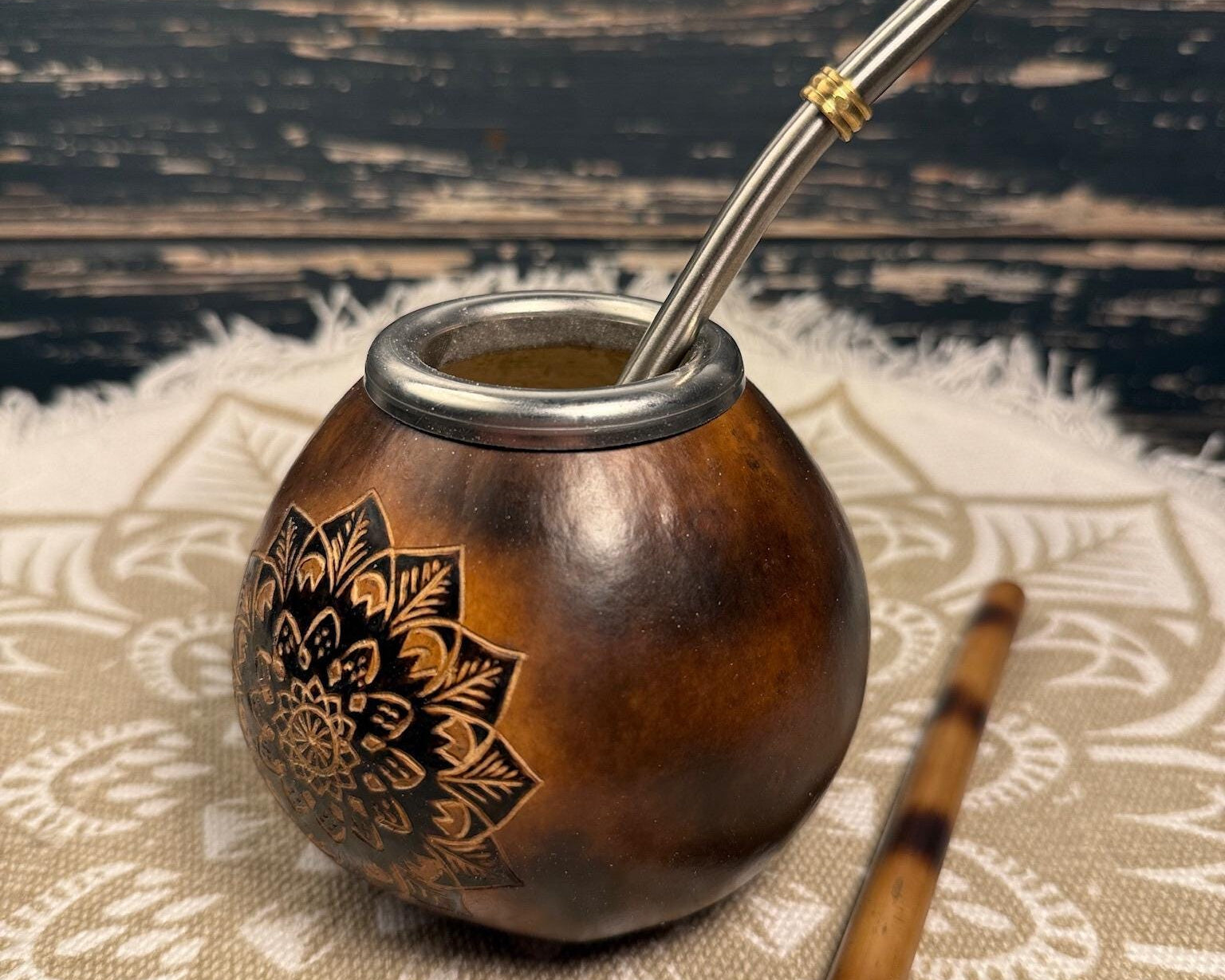 A brown wooden mate bowl with a black floral mandala design on the side, filled with mate tea and topped with a silver metal straw. A wooden spoon rests beside it on a white patterned cloth over a dark rustic wooden background.
